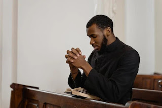 A man in a black robe is praying solemnly inside a church, surrounded by stained glass windows and wooden pews.